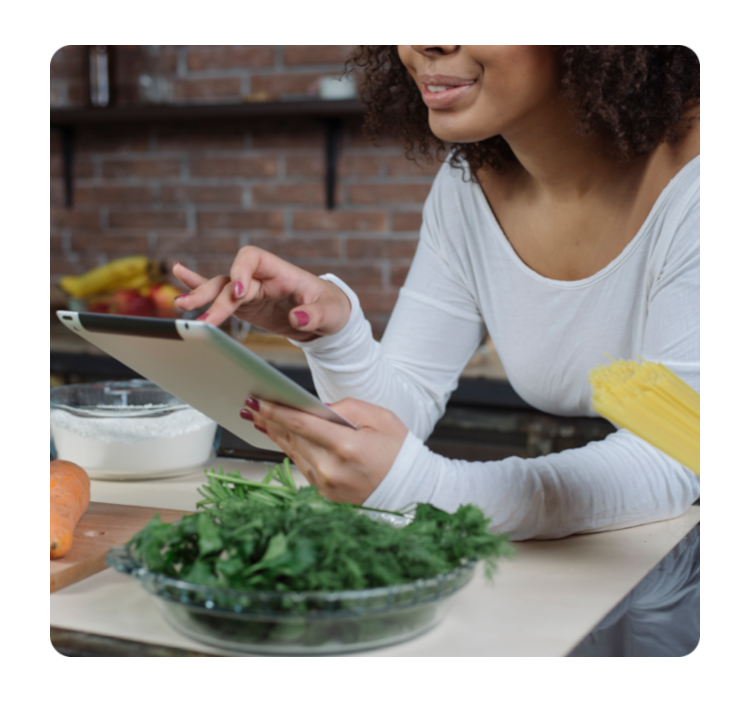 Persona utilizando una tablet en la cocina, rodeada de ingredientes frescos como vegetales y harina, mientras prepara una receta.