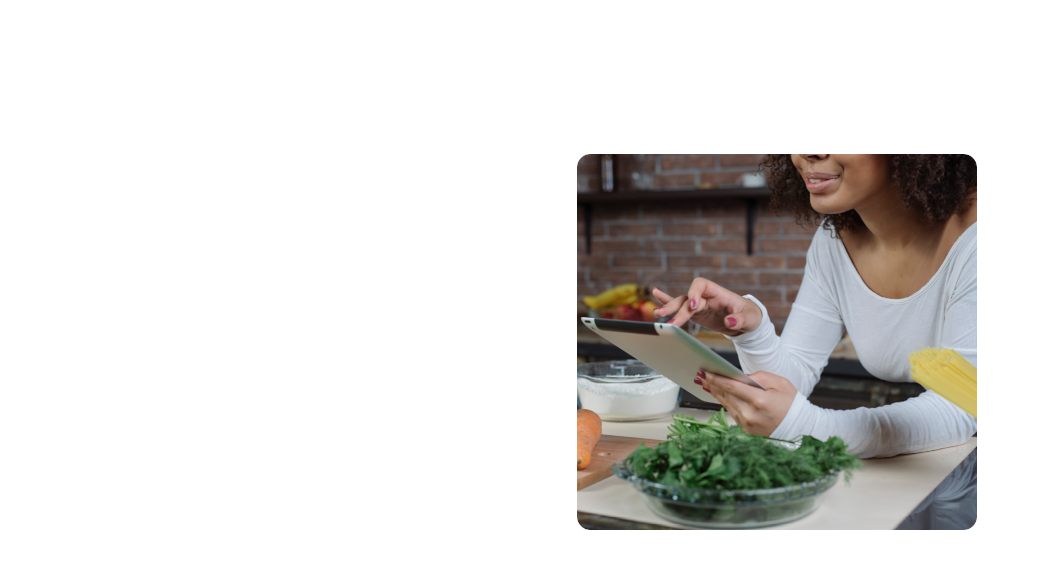 Persona utilizando una tablet en la cocina, rodeada de ingredientes frescos como vegetales y harina, mientras prepara una receta.